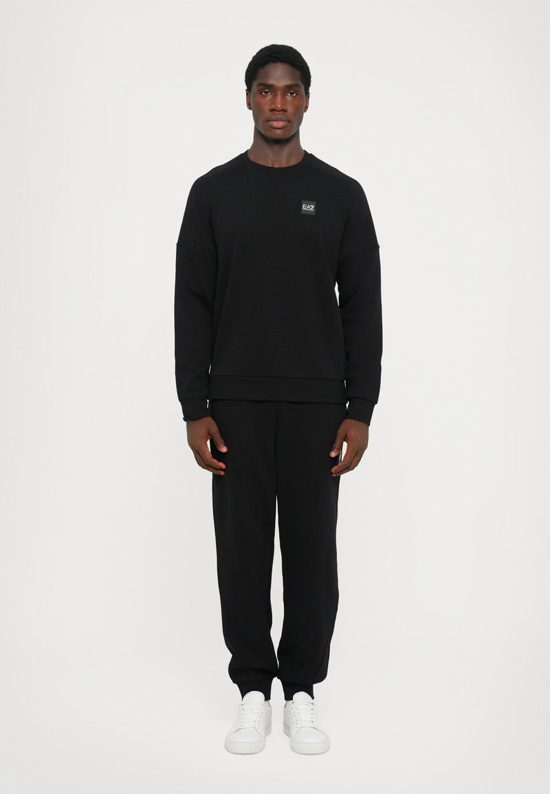 Felpa nera con scollo tondo e patch etichetta, abbinata a pantaloni neri coordinati. Indossata con scarpe da ginnastica bianche, su uno sfondo semplice.