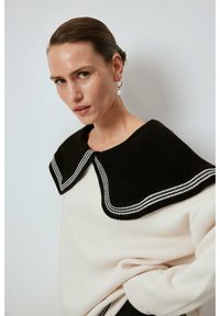Maglione a maglia con colletto bianco e nero, caratterizzato da un corpo crema. Il colletto presenta una texture a coste e strisce sportive lungo il bordo.