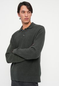 Pullover verde scuro, strutturato, con colletto, apertura con bottoni e maniche lunghe. Il tessuto sembra morbido e versatile.