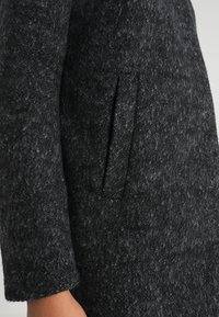 Cappotto di lana grigio scuro con superficie texture, tasca laterale e maniche ampie, con silhouette aderente e linee pulite.
