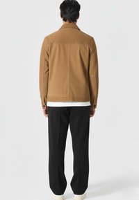 Brązowa kurtka z korduroju z klasycznym kołnierzem, zapinanymi mankietami i prostym krojem, zestawiona z czarnymi spodniami i białymi butami.