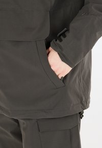 Mørk grå vandtæt jakke med lomme i siden, der lukkes med lynlås, og justerbare ærmer med velcro-detaljer. Glat, mat tekstur.