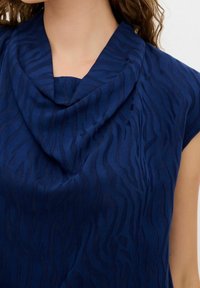 Top blu navy con scollo a cappuccio, caratterizzato da un motivo testurizzato e ondulato. Tessuto morbido con maniche corte e leggero effetto lucido.