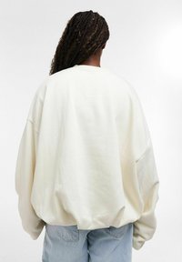 Personne aux cheveux tressés portant un sweat-shirt blanc surdimensionné et un jean bleu clair, debout face à un fond blanc uni.