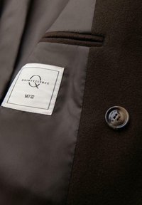 Binnenkant van een bruine wolmix jas met een grijze voering, voorzien van een label met het "Quintessence" logo en een enkele ronde knop.