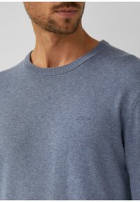 Mann trägt einen hellblauen Rundhals-Pullover mit dezentem gesticktem Logo auf der Brust, zeigt den Hals und einen Teil des Gesichts mit Stoppeln.