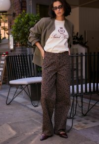 Oliven grøn oversized jakke, hvid t-shirt med logo, brune leopard mønstrede vidde bukser og bordeaux spidse flade sko mod en urban baggrund.