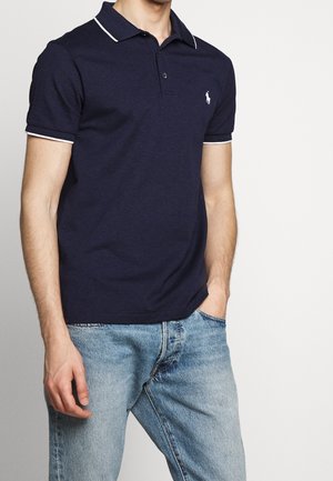 Homme portant un polo bleu marine avec un liseré blanc sur le col et les manches, associé à un jean bleu clair, debout avec une main dans la poche.