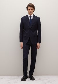 Traje azul marino con rayas finas, diseño entallado, pantalones a juego y camisa blanca. Completado con una corbata oscura y zapatos de cuero negros.
