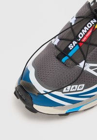 Sapatilha de corrida cinza e azul com parte superior em malha, detalhes texturizados, atacadores planos e acabamentos brancos. Possui sola em borracha e marcação lateral.