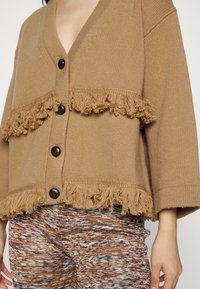 Cardigan marron en tricot avec ourlet à franges et fermeture à boutons. Présente des manches larges et un tissu texturé. Associé à un pantalon multicolore à motifs.