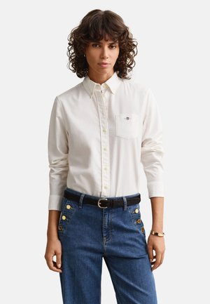 Frau mit lockigem Haar, die ein weißes, geknöpftes Hemd trägt, das in blaue hoch taillierte Jeans mit goldenen Knöpfen und einem schwarzen Gürtel gesteckt ist.