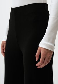 Pantalons noirs côtelés à taille haute, avec une texture lisse et un design à jambes larges. Une chemise blanche à manches longues est partiellement visible.