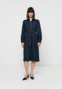 Denim krekls-duksa ar garajām piedurknēm, jostasvietas akcentu un vidējo garumu. Iezīmējas ar gaiši zilu denimu un izšūtu detaļu visā virsmā.