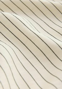 Tissu en coton beige avec de fines rayures verticales noires, doté d'une texture lisse et d'un léger éclat. Convient à divers modèles de vêtement.