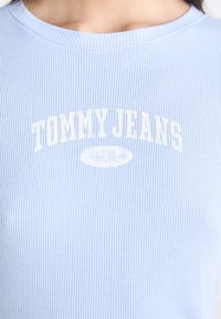 Gaiši zilā ribota T-kreklu ar „TOMMY JEANS” zīmolu baltā krāsā virs apaļā „Jaunā Jorkas” logo. Mīksta tekstūra ar apaļu kakla izgriezumu.