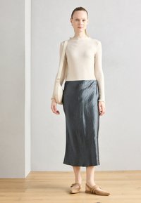 Top beige a maniche lunghe a coste con scollatura a volant, abbinato a una gonna a tubino in satin grigio scuro e scarpe basse marrone chiaro con dettagli.