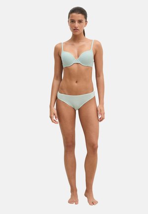 Vrouw staat blootsvoets, gezicht naar voren, draagt een lichtgroen gestreepte beha en bijpassende bikinitrukjes tegen een effen lichte achtergrond.
