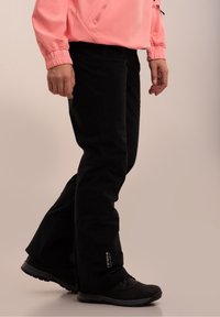 Personne portant un pantalon de neige noir et des chaussures noires avec une veste rose visible de la taille vers le bas sur un fond uni.