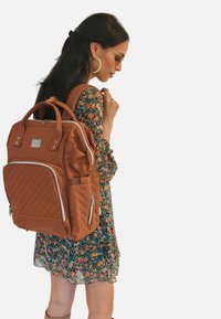 Mochila acolchada marrón con dobles cremalleras, bolsillo lateral y asas en la parte superior. El material parece liso y duradero, con detalles en plateado.