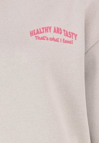 Hellgrauer Pullover mit gerippter Textur, versehen mit einem rosafarbenen gestickten Schriftzug, der "GESUND UND LECKER Das ist, was ich liebe!" sagt.