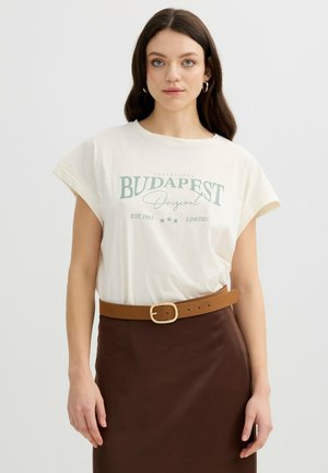 Mujer con cabello largo y oscuro, lleva pendientes de aro dorados y una camiseta crema con la palabra "Budapest" metida dentro de una falda marrón con cinturón beige sobre un fondo liso.