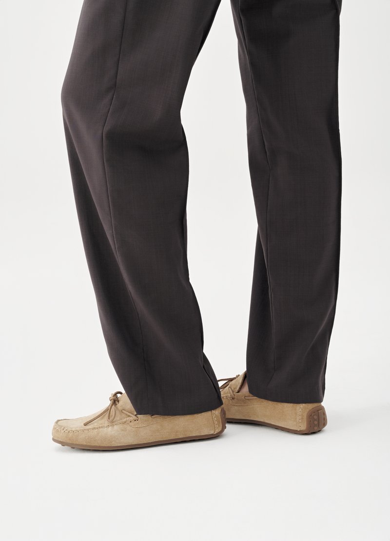 Close-up van benen die donkere broek en lichtbruine suède loafers met rubberen zolen dragen, op een witte achtergrond.