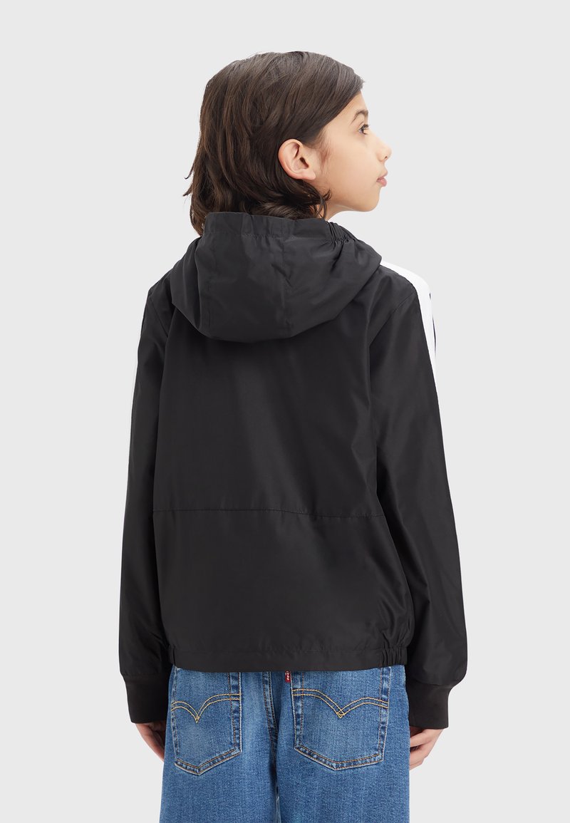Kind mit schulterlangen dunklen Haaren, das eine schwarze Kapuzenjacke und blaue Jeans trägt, seitlich vor einem schlichten Hintergrund stehend.