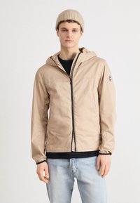 Lightweight beige jacket with a hood, full zipper, and black accents. Worn over a black shirt and paired with light blue jeans.