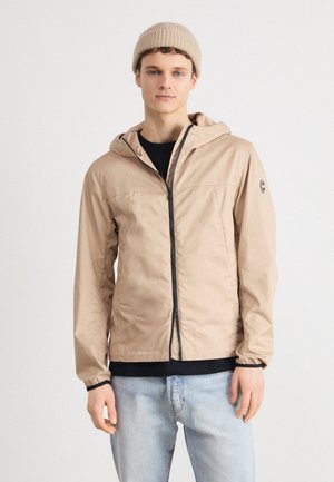 Giacca beige leggera con cappuccio, cerniera completa e dettagli neri. Indossata sopra una maglietta nera e abbinata a jeans azzurri chiari.