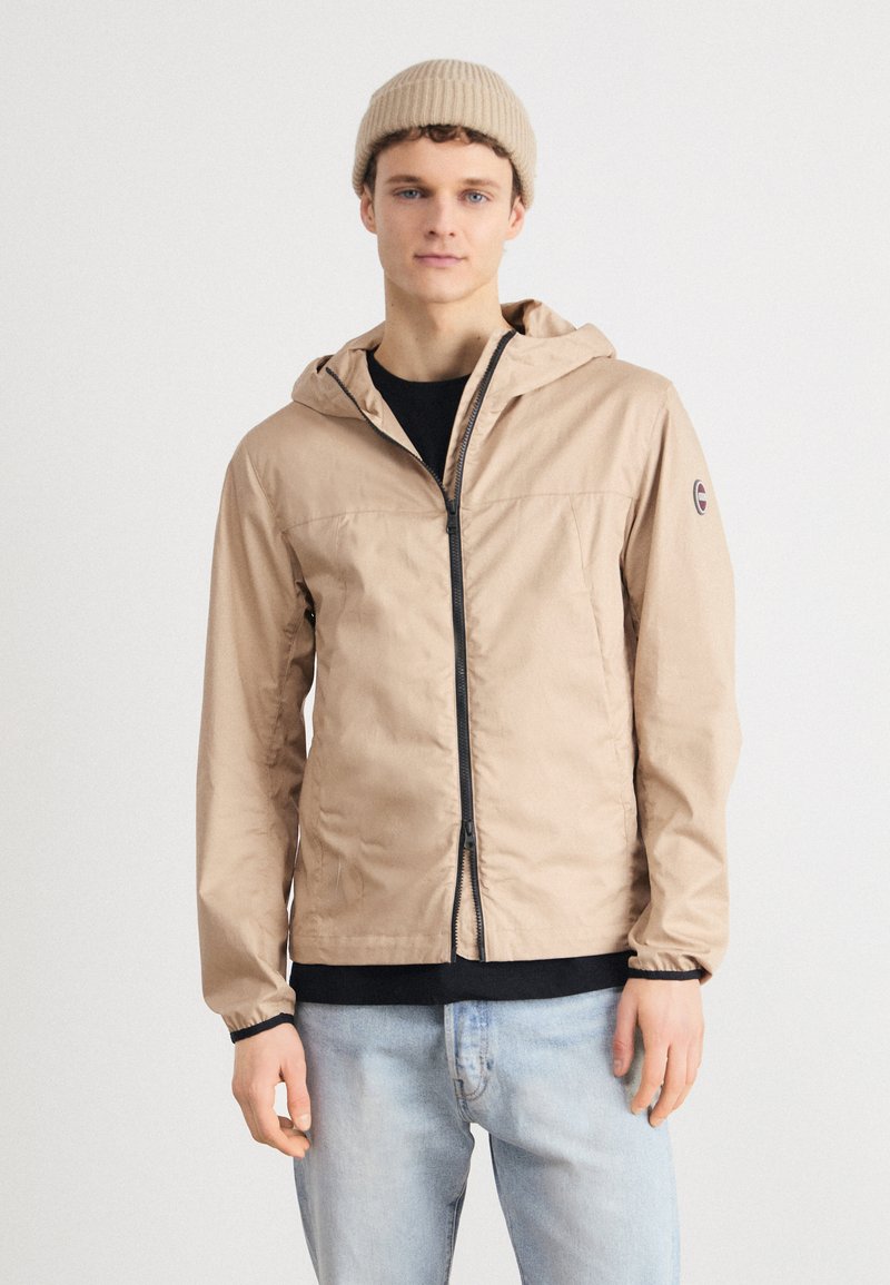 Lightweight beige jacket with a hood, full zipper, and black accents. Worn over a black shirt and paired with light blue jeans.