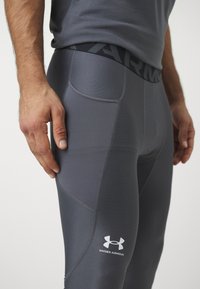 Szürke kompressziós leggings sima textúrával, magas derékkal, az "ARMOUR" felirattal és az Under Armour logóval az alsó lábon.