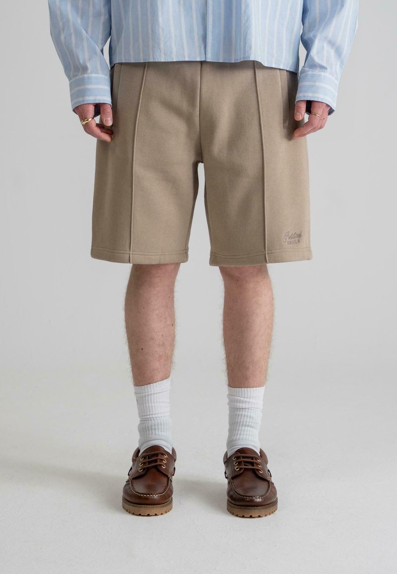 Beige shorts with seam detailing, made of soft fabric. Paired with brown shoes and white socks, worn with a light blue striped shirt.