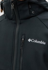 Veste softshell noire avec une poche poitrine zippée, une fermeture éclair complète sur le devant et une capuche ajustable avec cordons de serrage.