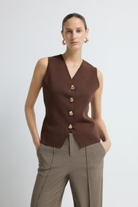 Donna che indossa un top senza maniche marrone con bottoni e dettagli dorati, abbinato a pantaloni a quadri a vita alta, in piedi con le mani in tasca contro un muro bianco.