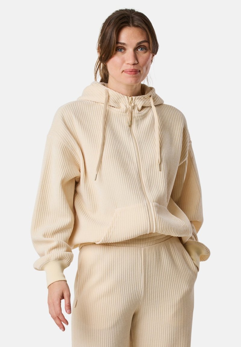 Felpa a costine color crema con zip frontale, cappuccio grande e lacci. Presenta una tasca a marsupio e una vestibilità rilassata. Abbinata a pantaloni a costine.