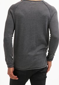 Chemise grise à manches longues avec un tissu texturé, dotée de manches raglan et de coutures contrastées le long des seams, portée avec un pantalon noir.