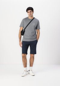 Herren grå bomull t-skjorte med logo på brystet, kombinert med marineblå shorts. Brukt med en svart crossbody-veske og hvite joggesko, stående mot en hvit bakgrunn.