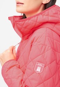 Chaqueta de coral acolchada con un alto cuello y cierre de cremallera. Presenta un parche con el logo en la manga izquierda y un tejido suave y texturizado.