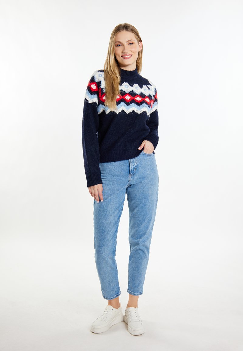 Marineblauer Strickpullover mit einem mehrfarbigen Zickzackmuster in Rot, Weiß und Hellblau. Kombiniert mit hellblauen Jeans und weißen Sneakers.