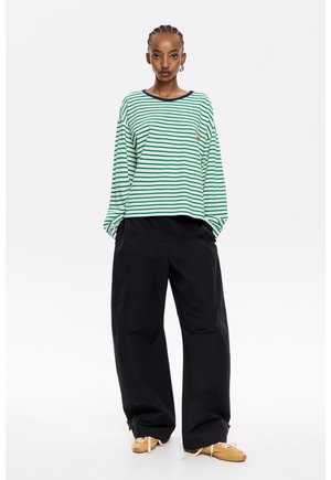 Grün-weiß gestreiftes Langarmshirt mit Rundhalsausschnitt, kombiniert mit lockeren schwarzen Hosen und hellbraunen Schuhen.