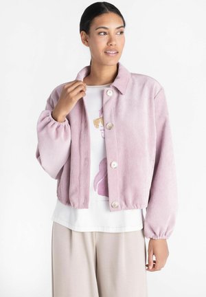 Vrouw draagt een lichtroze corduroy jas met knopen over een wit grafisch t-shirt en beige broek, kijkt naar de zijkant.