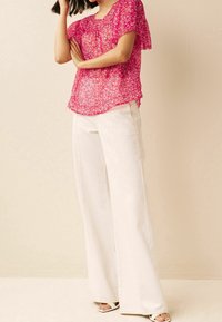 Blouse florale rose transparente à manches courtes, associée à un pantalon large blanc et des sandales blanches. Tissu simple et léger avec un design fluide.