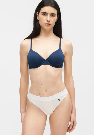 Γυναίκα με μακριά σκούρα μαλλιά που φοράει μπλε σουτιέν και λευκό μαγιό σλιπ, στέκεται μπροστά σε απλό φωτεινό φόντο.