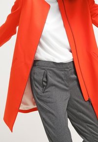 Světlý červený kabát s ostrým střihem, vyrobený z hladkého materiálu a s otevřeným předním dílem, kombinovaný se šedými proužkovanými kalhotami a bílým topem.