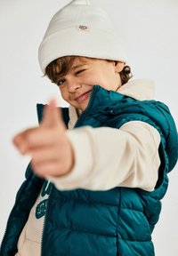 Un gilet rembourré en sarcelle avec fermeture éclair et poches latérales, superposé à un hoodie crème. Un bonnet en tricot blanc complète le look.