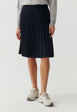 Person iført en knælængde sort plisseret nederdel, lysegrå sweater og beige sneakers, stående på en ensfarvet hvid baggrund.