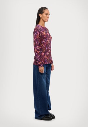 Langemouwige paarse top met abstracte spiraalpatroon, gecombineerd met losse blauwe denim jeans en zwarte plateauschoenen. Zijaanzicht.