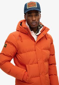 Orange puffer-jakke med en tekstureret finish, justerbar hætte og sidelommer. Båret over en ternet skjorte med hvid krave.