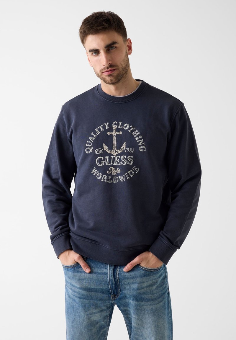 Homme portant un sweat-shirt bleu marine avec une ancre et le texte "Quality Clothing Guess Worldwide", associé à un jean bleu, debout devant un fond uni.